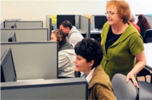 An instructor stands behind a student sitting at a computer workstation.
